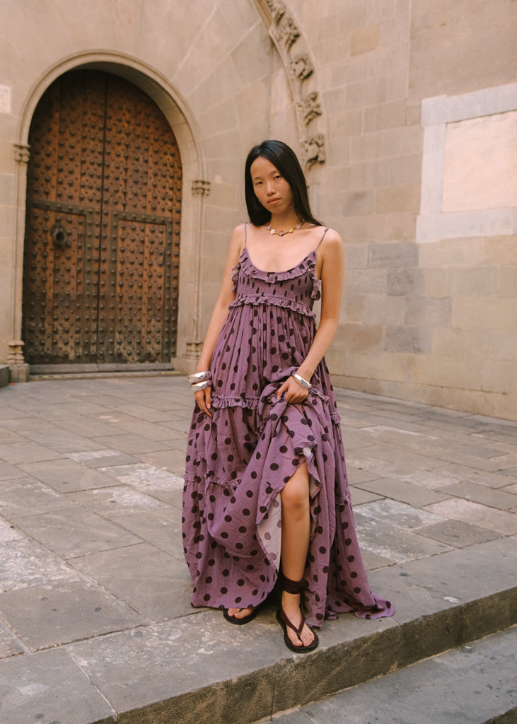 A woman wearing a purple maxi dress with dark brown polka dots stands on stone steps in front of a stone building with a large wooden door.