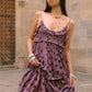 A woman wearing a purple and dark brown polka dot ruffled maxi dress, standing in front of a stone archway.