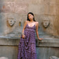 A woman wearing a purple tiered maxi dress with dark brown polka dots stands in front of a stone wall with sculpted faces.