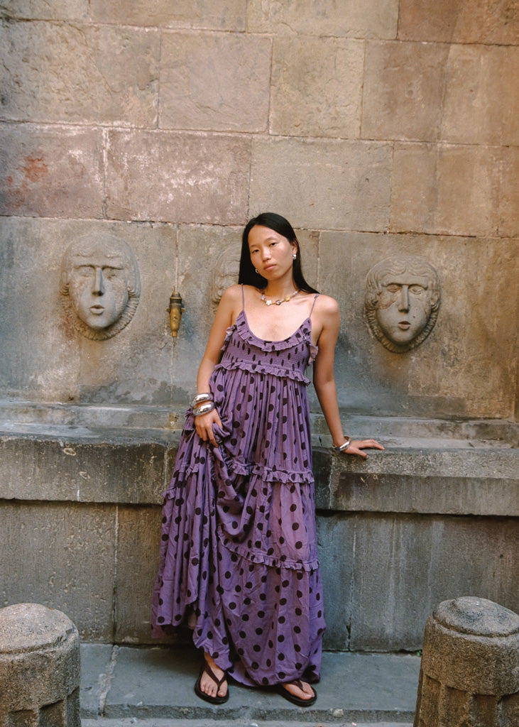 A woman wearing a purple tiered maxi dress with dark brown polka dots stands in front of a stone wall with sculpted faces.