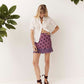 A woman modeling a white short-sleeve button-up shirt and a purple mini skirt with dark brown polka dots, standing against a white wall with green leaves in the corners.