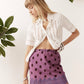 A woman modeling a white short-sleeve button-up shirt and a purple mini skirt with dark brown polka dots, standing against a white wall with green leaves in the corners.
