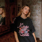A woman wearing a black oversized graphic t-shirt with pink 'Kinga Csilla' text and a floral design, standing in a vintage room with patterned wallpaper and a wooden mirror.