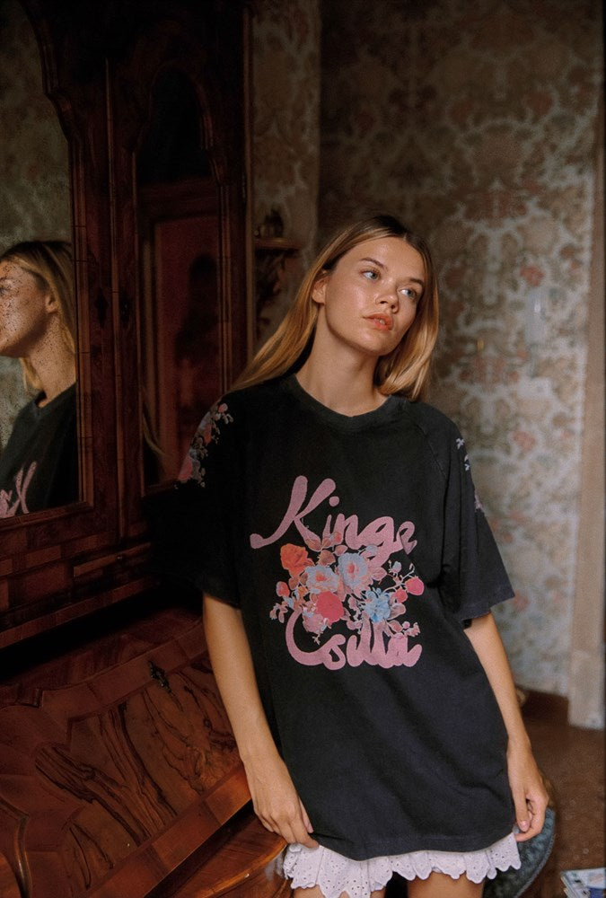 A woman wearing a black oversized graphic t-shirt with pink 'Kinga Csilla' text and a floral design, standing in a vintage room with patterned wallpaper and a wooden mirror.