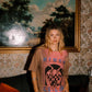 A woman stands in a vintage room wearing a faded pink graphic t-shirt with 'KINGA CSILLA' printed on the front, paired with blue and black patterned pants. Behind her is a large framed landscape painting and floral wallpaper.