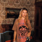 A woman stands in a vintage room with patterned wallpaper, wearing a brown graphic t-shirt with a pinkish-orange tint and dark patterned pants.