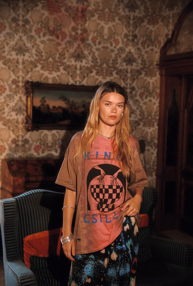 A woman stands in a vintage room with patterned wallpaper, wearing a brown graphic t-shirt with a pinkish-orange tint and dark patterned pants.