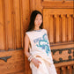 A woman stands against a rustic wooden door wearing a cream sleeveless top with a blue dragon graphic and the text 'KINGA CSILLA', paired with matching cream lace shorts.