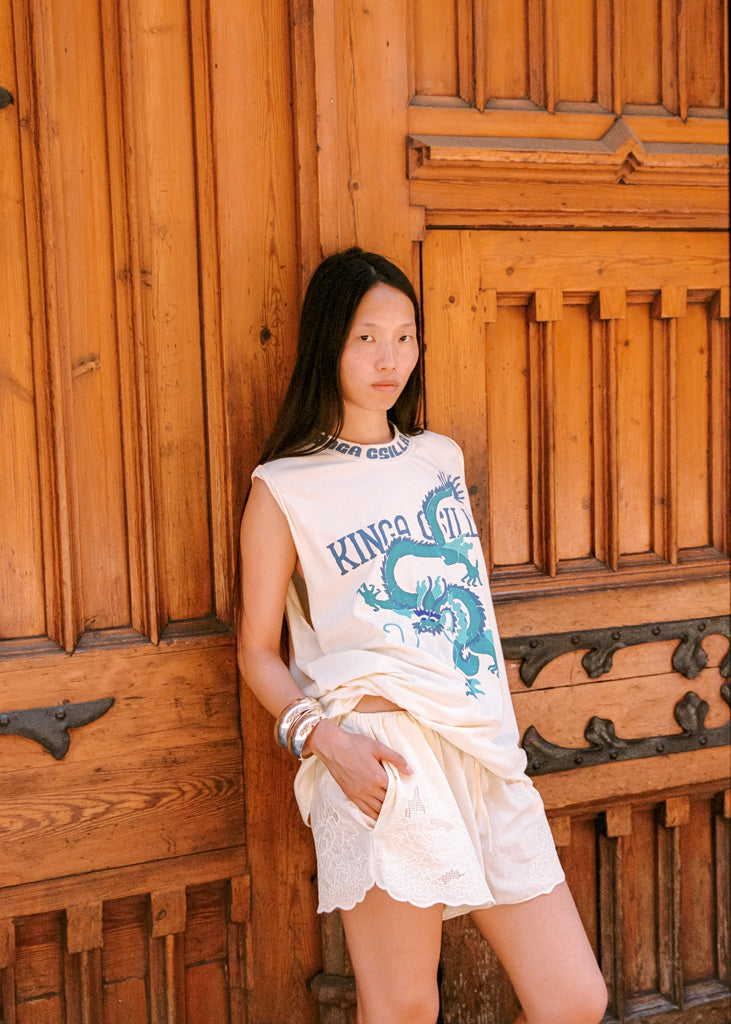 A woman stands against a rustic wooden door wearing a cream sleeveless top with a blue dragon graphic and the text 'KINGA CSILLA', paired with matching cream lace shorts.