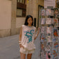 A woman stands on a city street wearing an oversized cream graphic T-shirt with a blue dragon print and the words 'KINGA CSILLA,' paired with cream shorts and black knee-high boots. She is posed beside a wire display rack filled with colorful postcards.