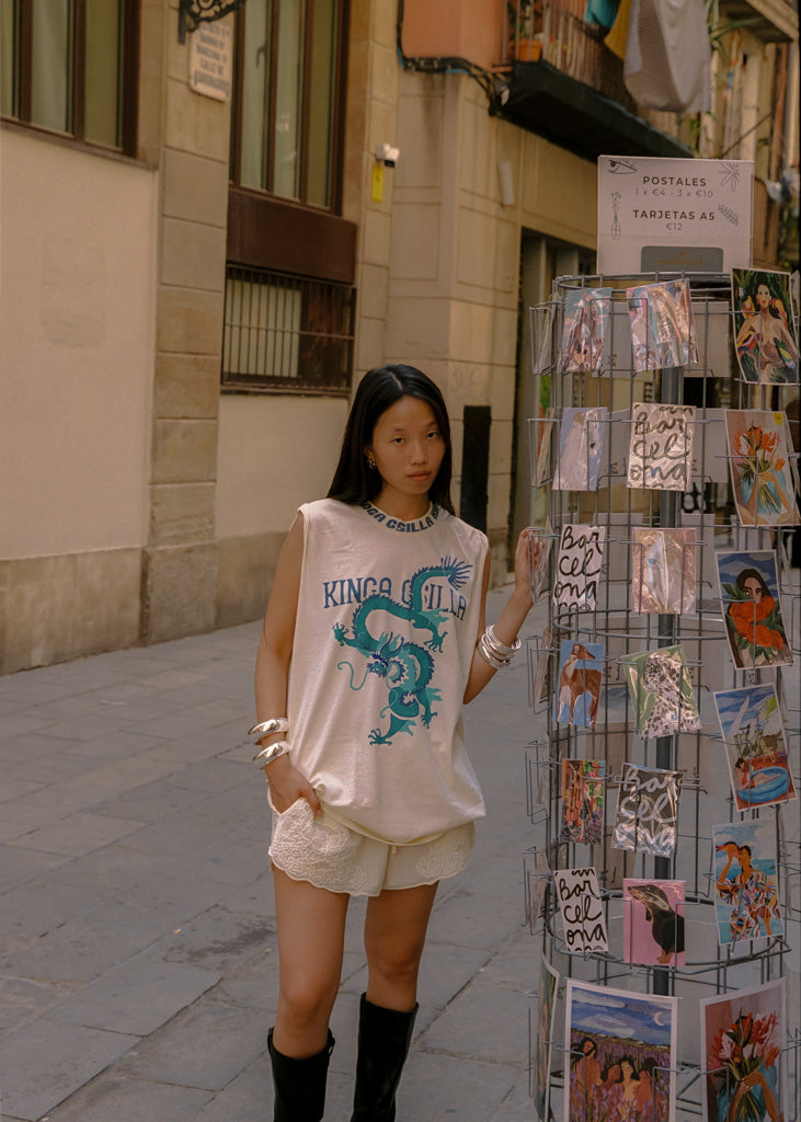 A woman stands on a city street wearing an oversized cream graphic T-shirt with a blue dragon print and the words 'KINGA CSILLA,' paired with cream shorts and black knee-high boots. She is posed beside a wire display rack filled with colorful postcards.