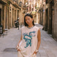 A woman stands on a narrow city street wearing a sleeveless cream top with a blue dragon graphic and the text 'KINGA CSILLA', paired with cream lace-trimmed shorts.