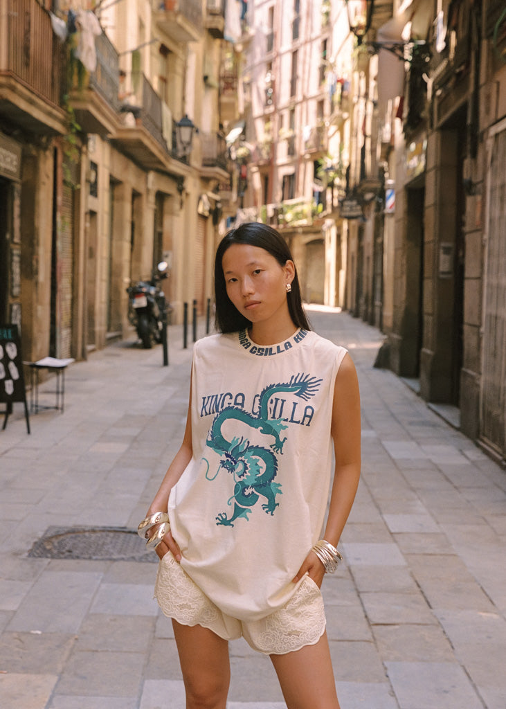 A woman stands on a narrow city street wearing a sleeveless cream top with a blue dragon graphic and the text 'KINGA CSILLA', paired with cream lace-trimmed shorts.