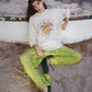 Person wearing a white sweatshirt with colorful design and green pants sitting on a log.