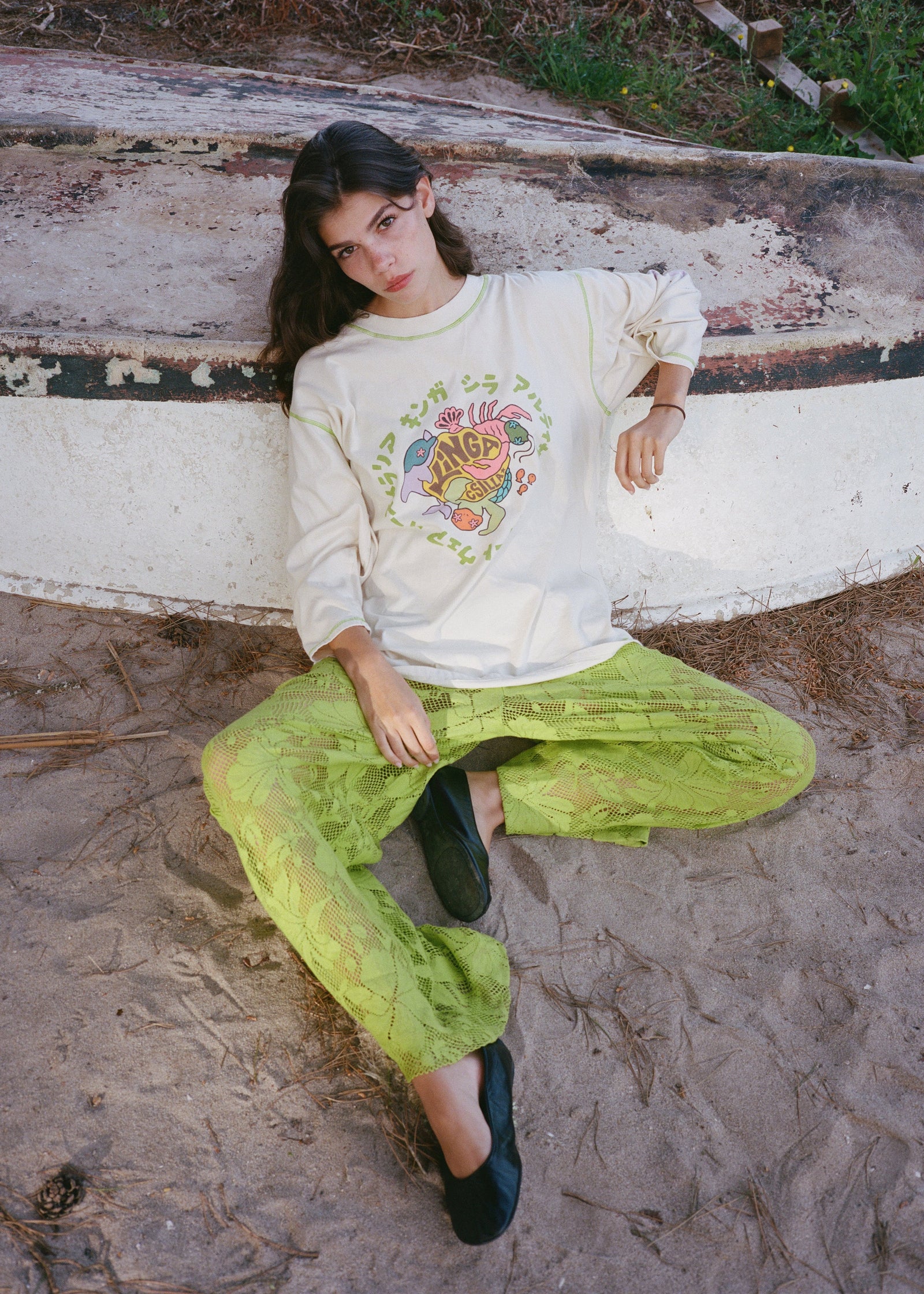Person wearing a white sweatshirt with colorful design and green pants sitting on a log.
