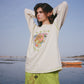 Woman wearing a white long-sleeve shirt with a colorful graphic and green pants standing by a body of water.