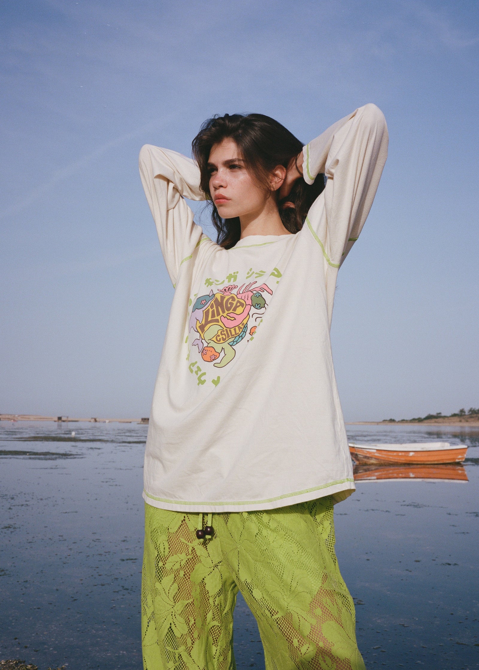 Woman wearing a white long-sleeve shirt with a colorful graphic and green pants standing by a body of water.