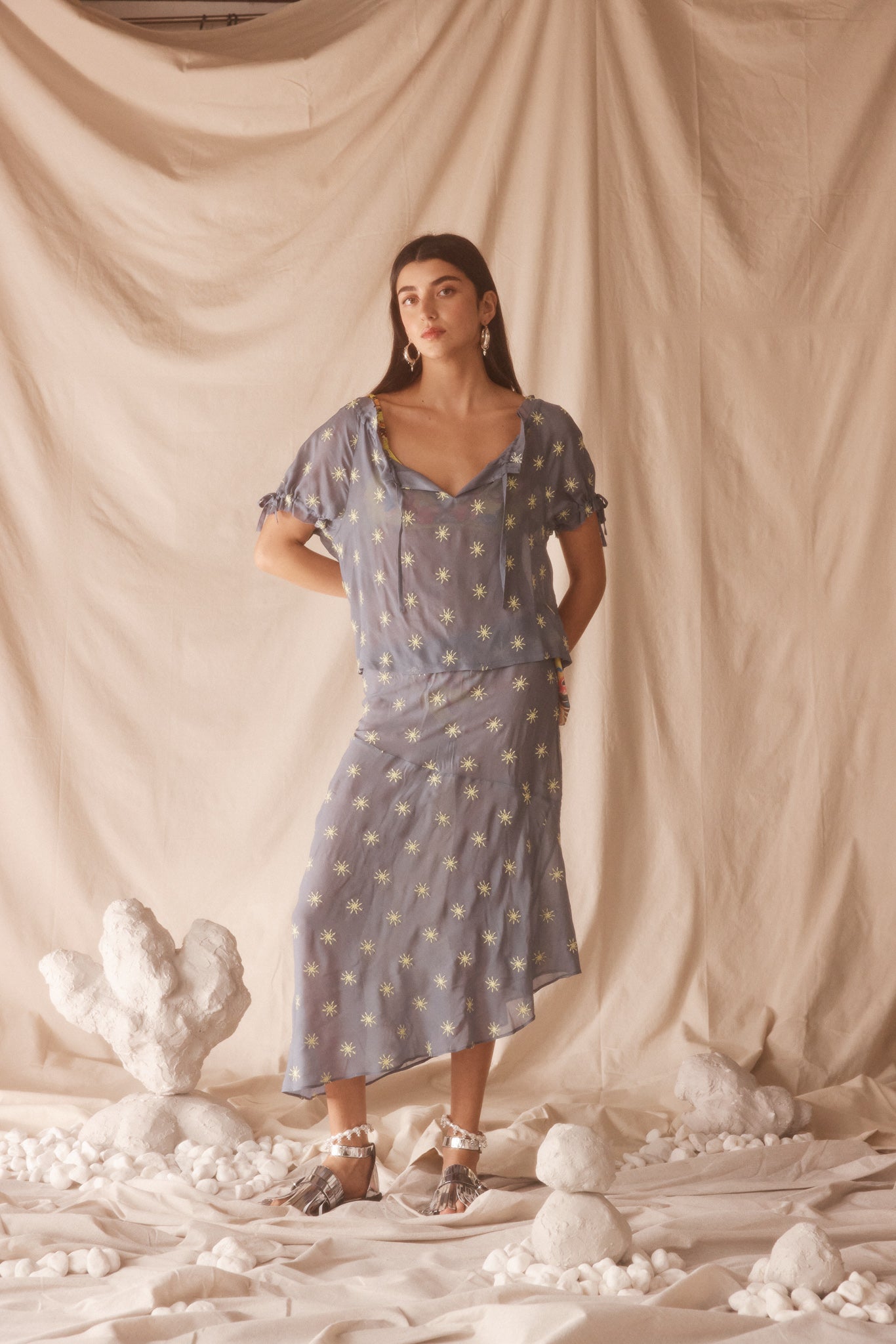 Woman in a sheer, blue blouse and midi skirt set with yellow sun embroided detailing. 