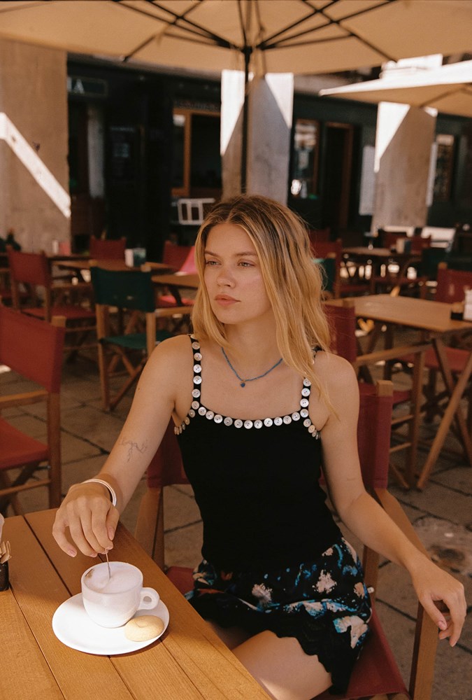 A woman sits at a wooden table at an outdoor cafe, wearing a black sleeveless top with white pearl-like embellishments along the neckline and a black floral skirt. She is holding a spoon over a white coffee cup on a saucer.