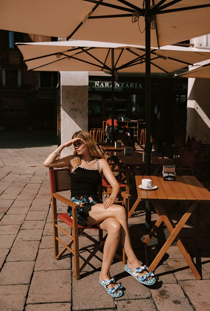 A woman sits on a wooden chair at an outdoor cafe table under a large umbrella, wearing a black top and floral shorts, with a cup on the table beside her.