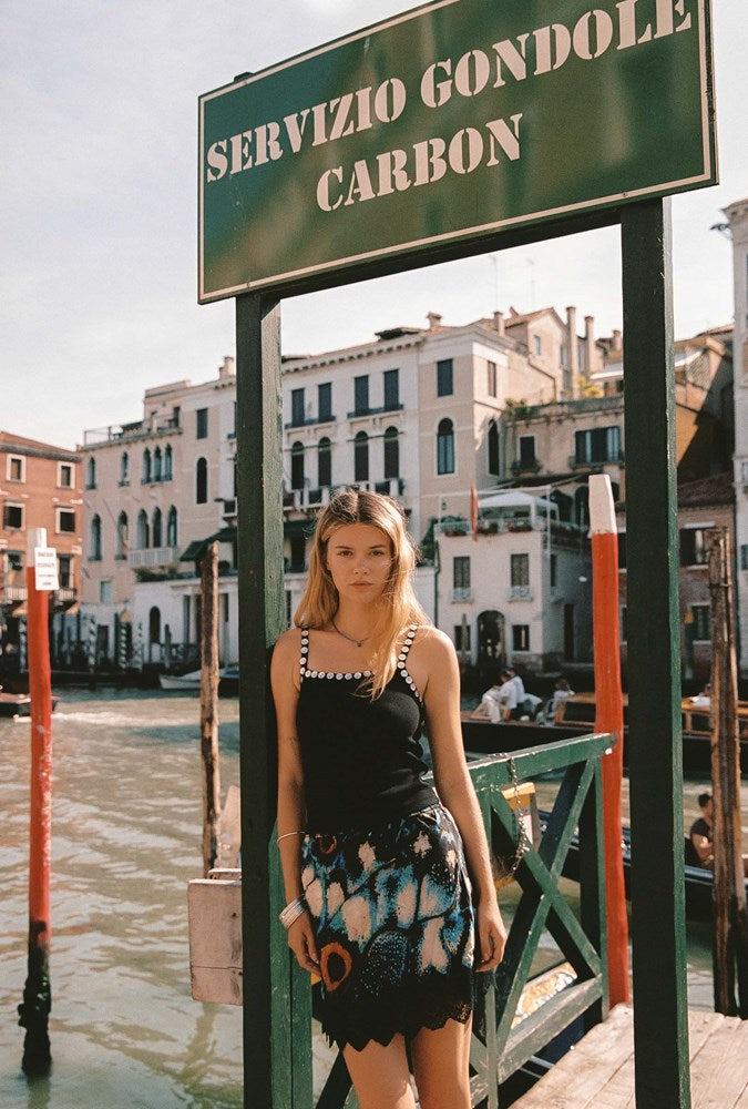 A woman stands on a wooden dock in Venice under a green sign reading 'SERVIZIO GONDOLE CARBON', wearing a black sleeveless top and a black, blue, and white patterned skirt. Water, canal buildings, and boats are visible in the background.