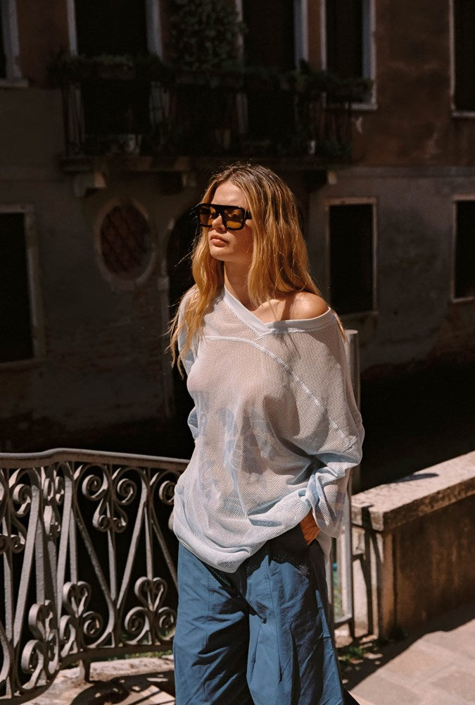 A woman wearing a sheer light blue off-the-shoulder top and blue wide-leg pants, standing outdoors in front of a building with a metal railing.