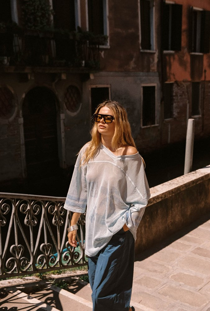 A woman wearing a sheer, light blue off-the-shoulder blouse and dark blue pants, standing on a stone walkway in an outdoor canal-side setting with historic buildings in the background.