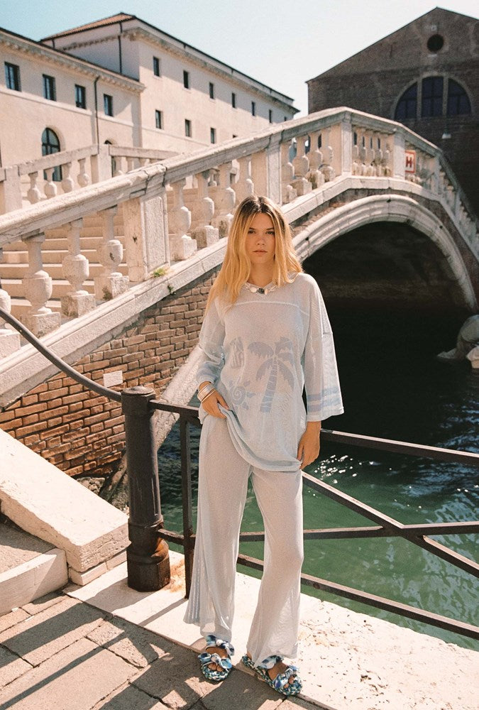 A woman stands on a stone bridge over water wearing a light blue and white patterned tunic top with matching wide-leg pants and patterned sandals, with an arched bridge and buildings in the background.