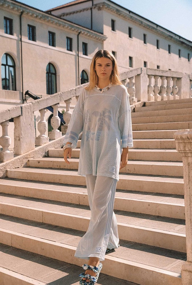 A woman walks down a large outdoor stone staircase wearing a loose, semi-sheer light blue long-sleeve tunic top and matching wide-leg trousers. The setting features a historic beige building in the background under bright daylight.