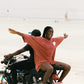 Two people on a motorcycle with one person in a red t-shirt dress, riding on a beach.