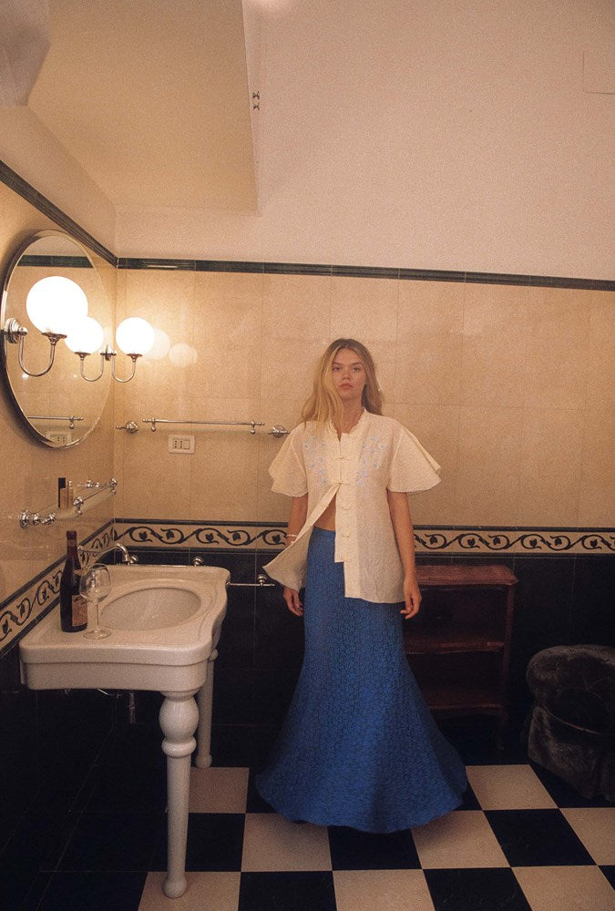 A woman stands in a vintage-style bathroom with a black and white checkered floor, a white sink, and tiled walls, wearing a cream puff-sleeve jacket over a blue textured maxi skirt.