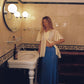 A woman stands in a vintage-style bathroom wearing a cream short-sleeve button-up shirt with ruffled sleeves and a long blue patterned maxi skirt. The bathroom features a white pedestal sink, a large round mirror with dual glowing globe lights, and a black-and-white checkered floor.