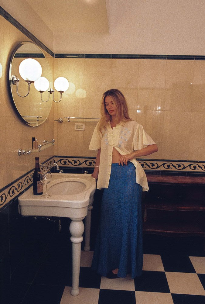 A woman stands in a vintage-style bathroom wearing a cream short-sleeve button-up shirt with ruffled sleeves and a long blue patterned maxi skirt. The bathroom features a white pedestal sink, a large round mirror with dual glowing globe lights, and a black-and-white checkered floor.