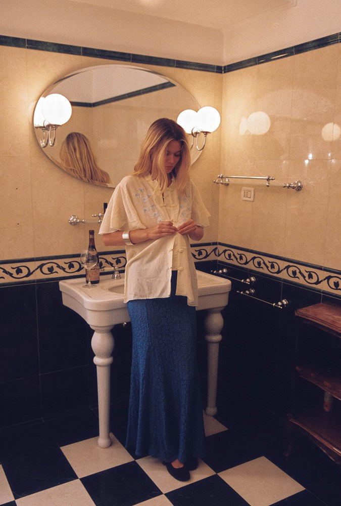 A woman stands in a vintage-style bathroom with black and white checkered flooring, beige tiled walls, and a white pedestal sink. She is wearing a cream-colored short-sleeve button-up shirt and a long blue skirt.