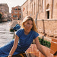 A woman wearing a textured blue short-sleeve dress leans on the polished wooden hull of a boat in a canal setting with historic brick buildings in Venice.