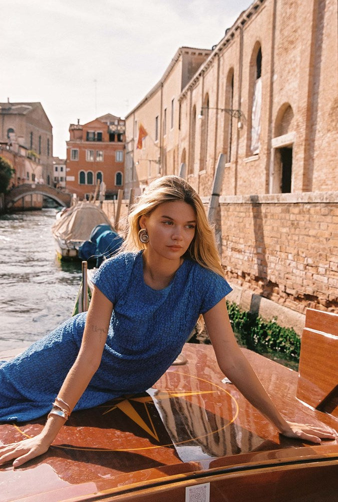 A woman wearing a textured blue short-sleeve dress leans on the polished wooden hull of a boat in a canal setting with historic brick buildings in Venice.