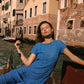 A woman wearing a textured blue short-sleeve dress reclines in a wooden boat on a canal with historic Venetian buildings in the background.