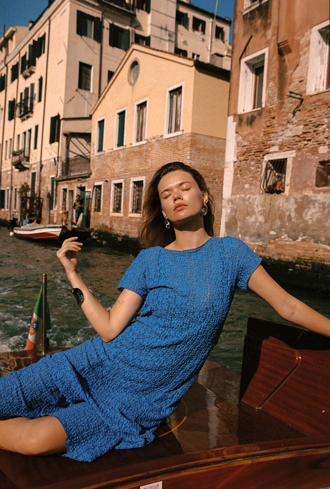 A woman wearing a textured blue short-sleeve dress reclines in a wooden boat on a canal with historic Venetian buildings in the background.
