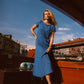A woman wearing a textured blue midi dress stands on the deck of a wooden boat with a canal and buildings in the background under a clear blue sky.