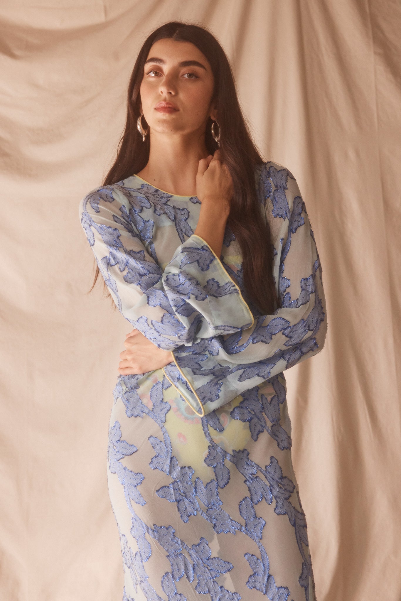 Woman wearing semi-sheer blue maxi with gold lurex florals, flared sleeves, and contrast piping. 