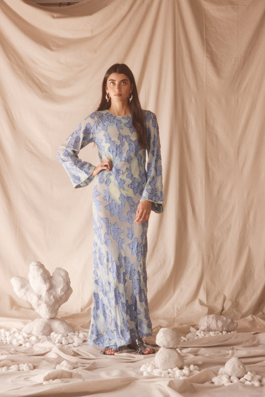 Woman wearing semi-sheer blue maxi with gold lurex florals, flared sleeves, and contrast piping. 
