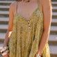 A woman wearing a sleeveless olive green and beige patterned slip dress with thin straps and a buttoned neckline, accessorized with chunky silver bracelets and a beaded necklace, standing outdoors against blurred stone steps.
