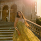 A woman wearing a voluminous olive green patterned gown stands on stone stairs in front of a grand classical building, with warm sunlight and lens flare creating a dreamy outdoor scene.