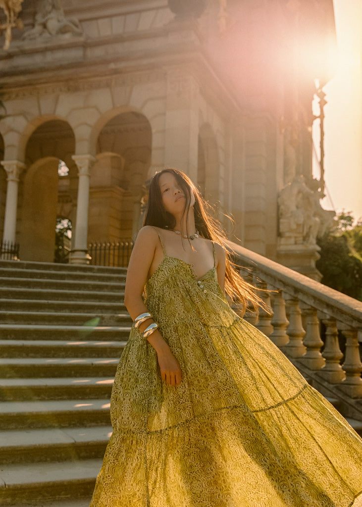 A woman wearing a voluminous olive green patterned gown stands on stone stairs in front of a grand classical building, with warm sunlight and lens flare creating a dreamy outdoor scene.