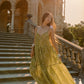 A woman wearing a flowing olive green patterned maxi dress walks down a grand stone staircase in front of a sunlit classical building.