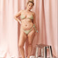 A woman models a light green and beige patterned bikini set in front of a pink curtain backdrop, with a small metallic stool to her right.