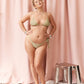 A woman modeling a light olive green bikini with a textured pattern, standing in front of a pink curtain backdrop with a small metallic stool to the side.