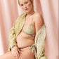 A woman seated on a metallic stool wearing an olive-green patterned bikini with a matching open-front cover-up draped off her shoulders, posed against a soft pink curtain backdrop.
