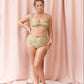 A woman models a two-piece bikini set with a textured, metallic gold and olive green pattern, standing against a draped pink fabric backdrop.