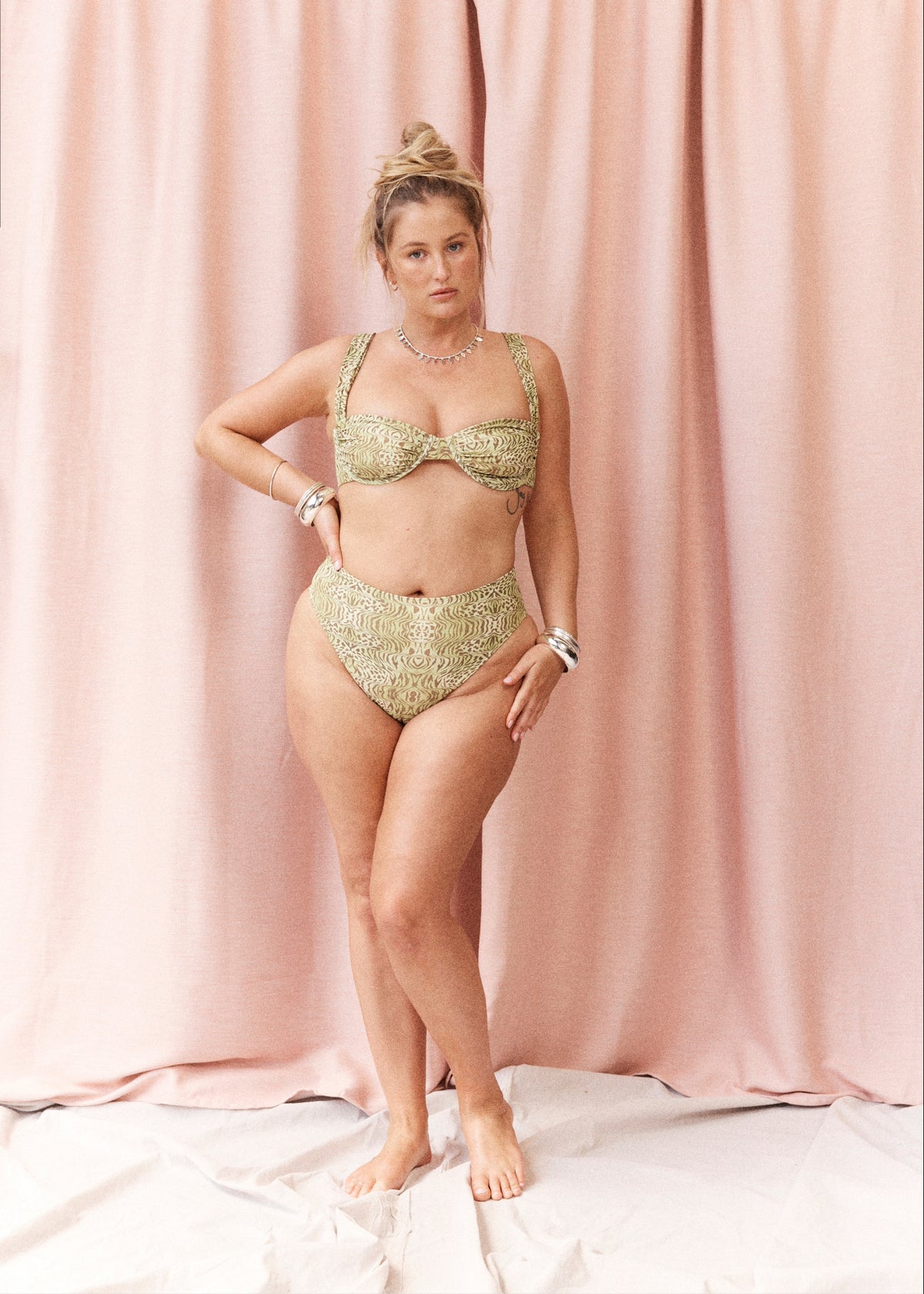 A woman models a two-piece bikini set with a textured, metallic gold and olive green pattern, standing against a draped pink fabric backdrop.